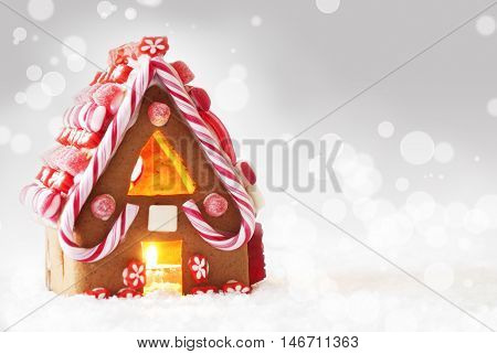 Gingerbread House In Snowy Scenery As Christmas Decoration. Candlelight For Romantic Atmosphere. Silver Background With Bokeh Effect. Copy Space For Advertisement