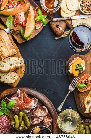 Italian antipasti wine snacks set. Brushettas, cheese variety, Mediterranean olives, pickles, Prosciutto di Parma with melon, salami and wine in glasses over black grunge background, top view, copy space