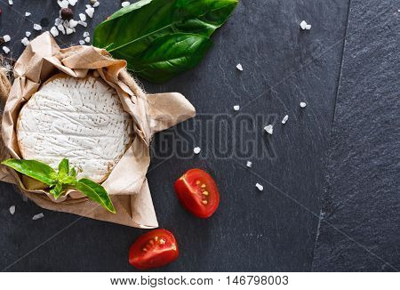 Cheese delikatessen closeup on black stone desk surface. Camembert or brie circle in brown kraft paper decorated with basil and pieces of cherry tomatoes, top view image with copy space