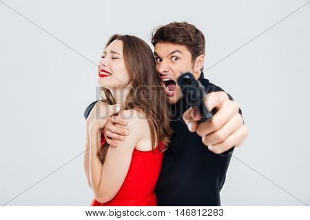 Furious criminal young man holding scared young woman and pointing with gun on you