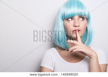 Closeup portrait of cheeky young girl in modern futuristic style wearing blue wig with curious expression and finger on lip while looking sideways over white wall background with copyspace