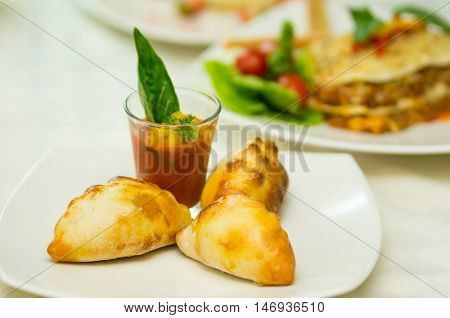 Apetizer of small tomato soup in shotglass and delicious mini empanadas spread around on white plate.