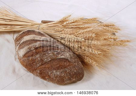 bunch of mown wheat ears with vintage handmade reaper hook sickle and rye dark french fresh bread loaf on white linen tablecloth