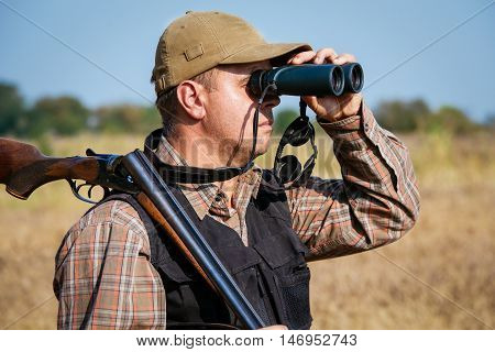 Man hunter with shotgun looking through binoculars in forest