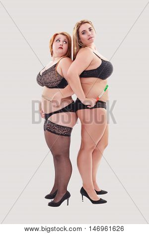 Two young chubby women in lingerie standing behind the other and have wrapped a tape measure around your body.