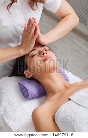 Head and face pummeling massage in spa. Female beautician work in wellness center. Professional massagist make relax healthy procedure to beautiful indian girl in cosmetology cabinet or beauty parlor.