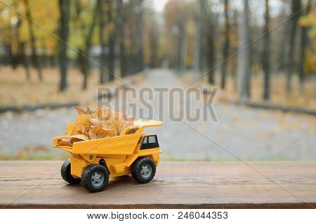 A Small Toy Yellow Truck Is Loaded With Yellow Fallen Leaves. The Car Stands On A Wooden Surface Aga