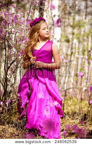 A Child With A Long Blond Hair Stands Among The Ledum And Birch In A Pink Dress And Smiling