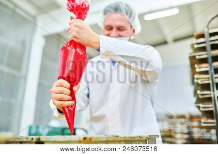Smiling Confectionery Factory Employee In White Coat Pouring Red Cream From Pastry Bag.