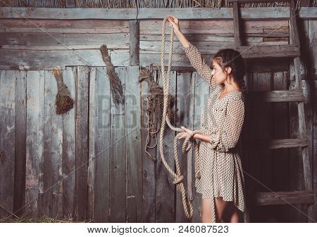 Young Girl In The Barn With Her Hand Up High Hanging A Piece Of Rope And Looking At It, Brooms And L