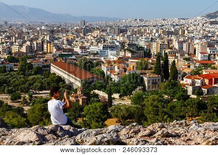 Toutist Sitting On A Rock Across The Acropolis Taking A Photo Of The Athens Cityscape On A Summer Af