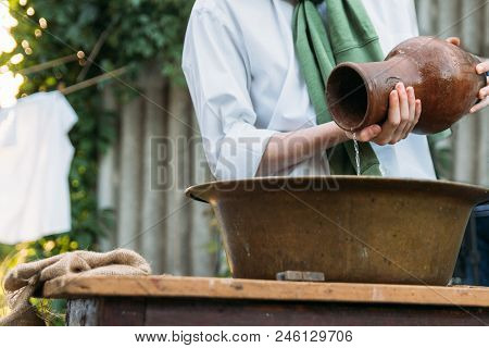 A Boy In A White Shirt And A Green Sweater, Holds A Clay Jug, Pours Water Out Of It Into A Copper Ba