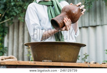 A Boy In A White Shirt And A Green Sweater, Holds A Clay Jug, Pours Water Out Of It Into A Copper Ba