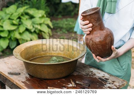A Boy In A White Shirt And A Green Sweater, Holds A Clay Jug, Pours Water Out Of It Into A Copper Ba