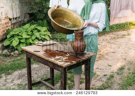 A Boy In A White Shirt And A Green Sweater, Holds A Clay Jug, Pours Water Out Of It Into A Copper Ba