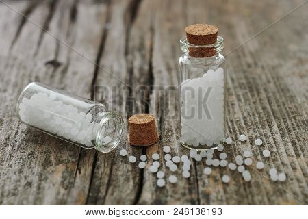 Granules Of Homeopathic Medicine In Small Glass Flasks And Scattered On An Old Wooden Table.