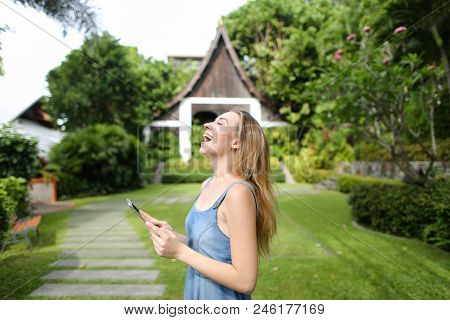 Young Pretty Girl Using Smartphone And Standing With Resort House In Background. Concept Of Modern T