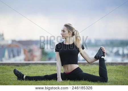 Happy Young Woman On The Grass Performs Twine With The Bent Leg, On Background Cityscape, Horizontal