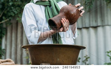 A Boy In A White Shirt And A Green Sweater, Holds A Clay Jug, Pours Water Out Of It Into A Copper Ba