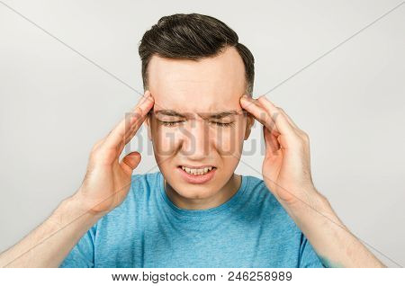 Young Guy With Headache Dressed In A Blue T-shirt On A Light Background.
