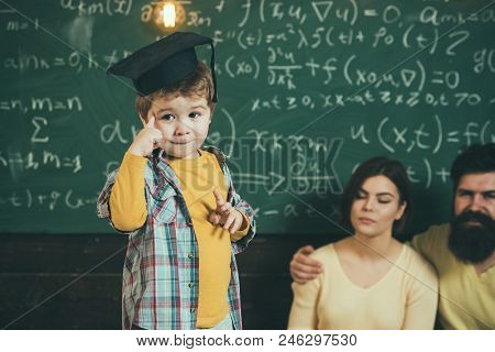 Smart Child, Wunderkind In Graduate Cap Pointing At His Head. Boy Presenting His Knowledge To Mom An