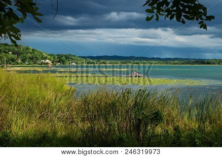 View On The Dock With People Along The Lake Shore With Dark Blue Cloudscape And Sunshine, El Remate,