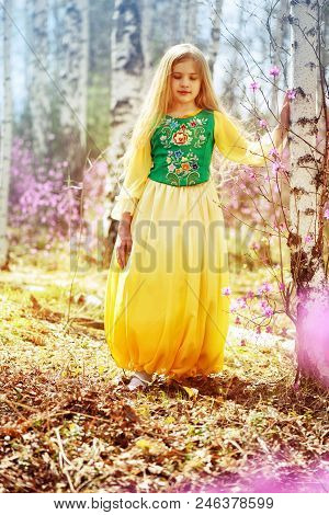 A Child With A Long Blond Hair Stands Among The Ledum And Birch In A Green Yellow Dress And Smiling