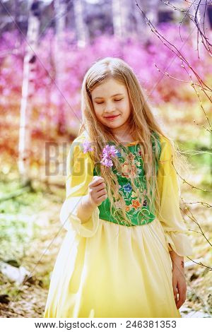 A Child With A Long Blond Hair Stands Among The Ledum And Birch In A Green Yellow Dress And Smiling