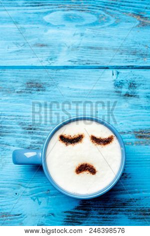 high-angle shot of a blue cup of cappuccino with a sad face drawn with cocoa powder on its milk foam, on a blue rustic table with a blank space on top