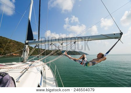 Young and happy sailor relaxes in a hammock set on the boom of the sailing boat anchored in a tropical sea