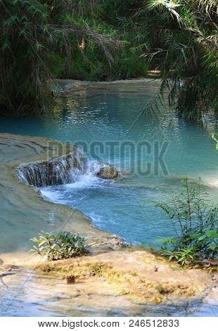 Part Of A River Has Formed A Pool Of Pale Blue Water. The Surrounding Forest Overhangs The Water. In