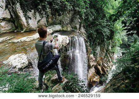A Traveler With A Phone Takes A Picture Of A Large Waterfall In The Agur Gorge On The Edge Of A Clif