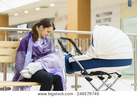 Young European Woman With Violet Stole Is Breastfeeding Her Little Child Close To White Baby Carriag