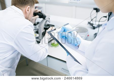 Crop Back View Of Female Microbiologist Writing Down Into Clipboard Data Received From Man Analyzing