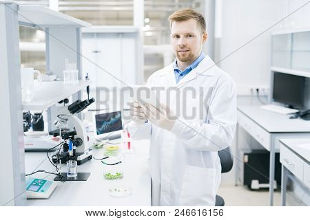 Young Male Microbiologist In Laboratory Coat Standing At Desk With Microscope And Green Vegetable Sa
