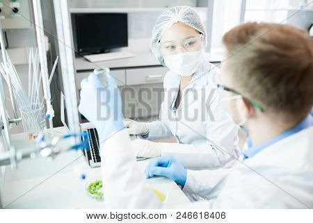 Microbiologists In Protective Laboratory Clothes Sitting At Table And Looking At Each Other Discussi