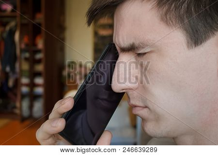 A Thoughtful Guy With A Smartphone At His Mouth, With His Eyes Closed, Sits At The Desk