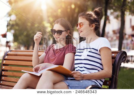 Outside Shot Of Cheerful Female Friends, Have Gentle Smiles, Prepare For Final Exam Together, Sit On