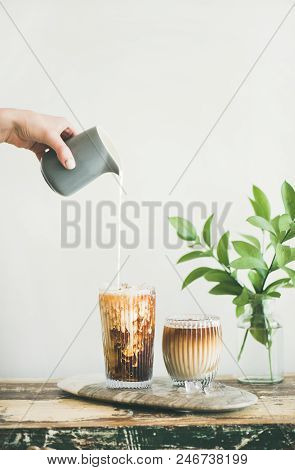 Iced Coffee In Tall Glasses With Milk Poured Over From Pitcher By Hand, White Wall And Green Plant B