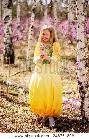 A Child With A Long Blond Hair Stands Among The Ledum And Birch In A Green Yellow Dress And Smiling
