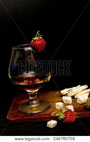Still Life With Cognac And French Blue Cheese On A Wooden Board. Glass With Cognac, Cheese, Strawber