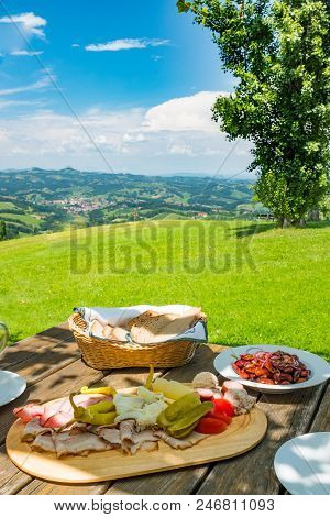 Brettljause With Cold Cuts, Beans Salad And Bread On Wooden Table With Beautiful View Over Green Mea