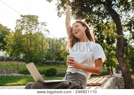 Image of shappy modern woman sitting on bench in green park and enjoying summertime while using silver laptop and drinking takeaway coffee from paper cup