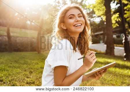 Photo of joyful woman 20s with broad smile sitting on green grass in park with legs crossed during summer day and writing down notes with pen