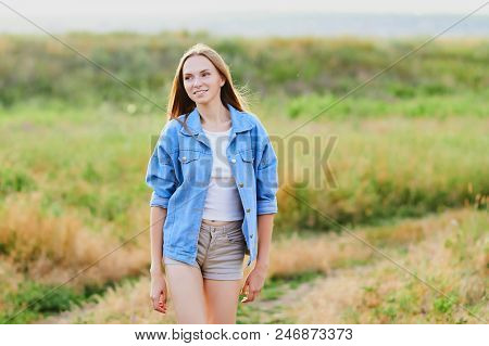 Happy Smiling Young Girl Wearing Blue Jeans Jacket And Shorts Is Standing In The Field