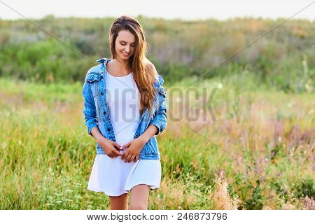Happy Smiling Young Girl Wearing Blue Jeans Jacket And White Dress Is Standing In The Field
