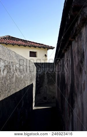 Narrow Pathway Between Dark Gray Stone Walls Of Matachin Fort In Sunlight, Baracoa, Cuba