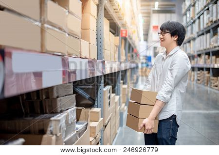 Young Asian Man Carrying Cardboard Boxes Near Product Shelf In Warehouse, Shopping Warehousing Or Wo