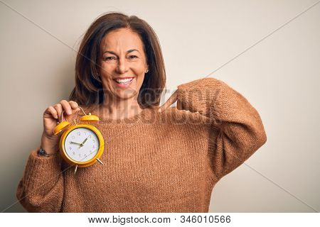 Middle age brunette woman holding clasic alarm clock over isolated background looking confident with smile on face, pointing oneself with fingers proud and happy.