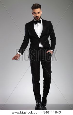Serious young model snapping his fingers and holding his hand in his pocket while wearing tuxedo, walking on gray studio background